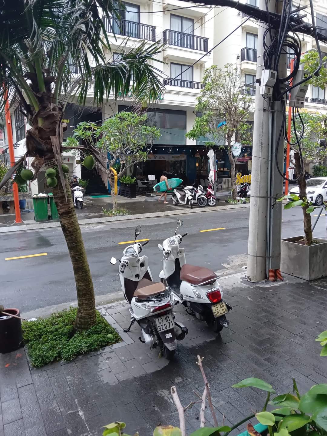 Surfer dude walking on the street to the beach, in Da Nang Vietnam. Two scooters are in the foreground on the sidewalk. Palm trees line the street