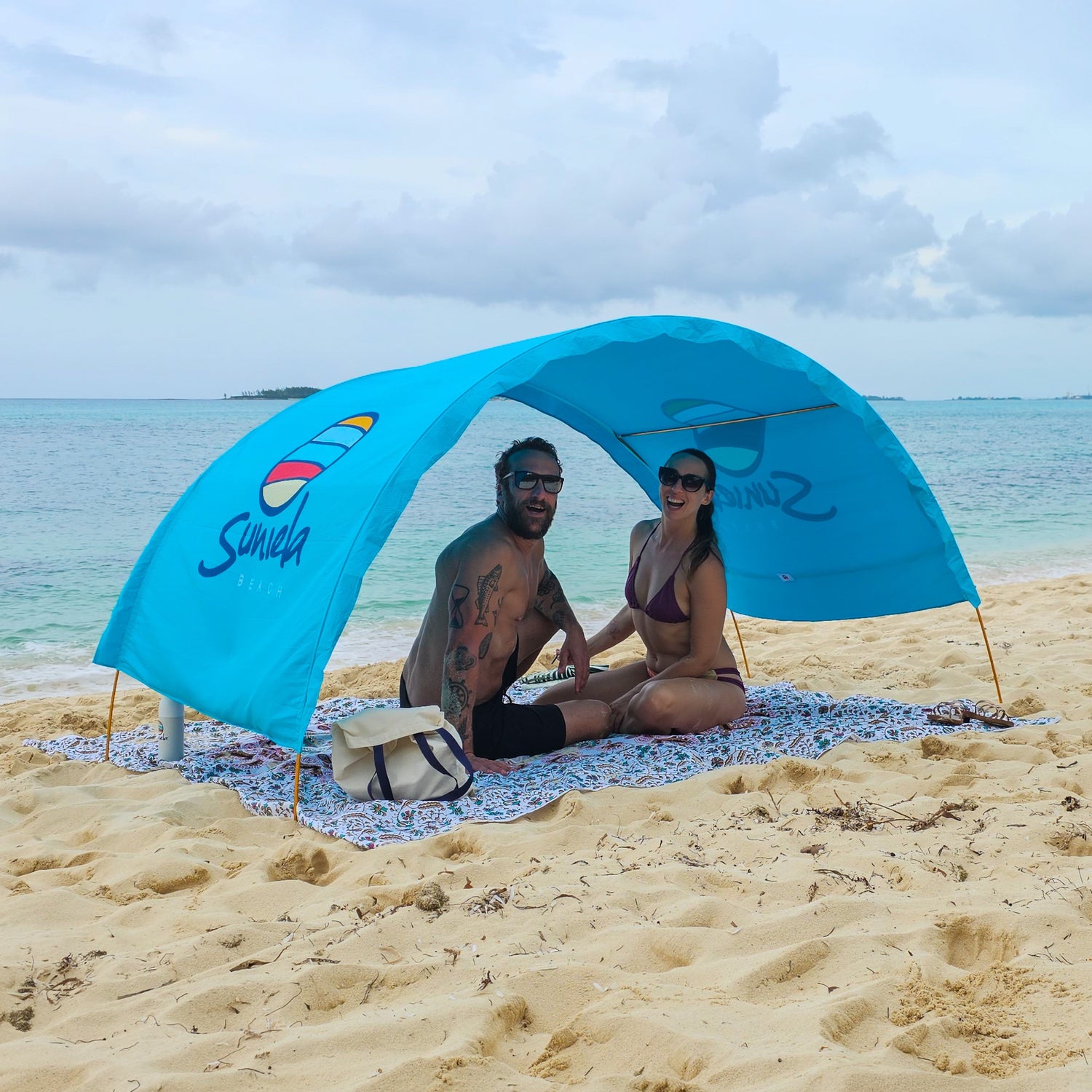 Couple sitting beneath an arched, blue Suniela shade cabana on a sandy beach