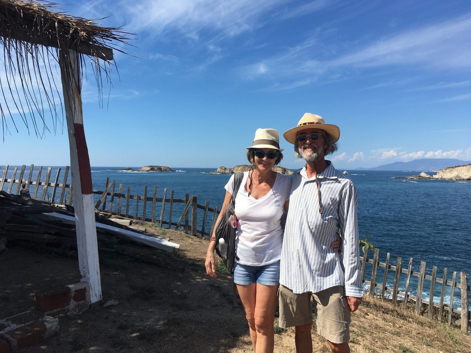 Chris and Marie-Christine travel the Michoacan Coast of Mexico, standing in front of a blue sea, wearing cool clothing and sun hats