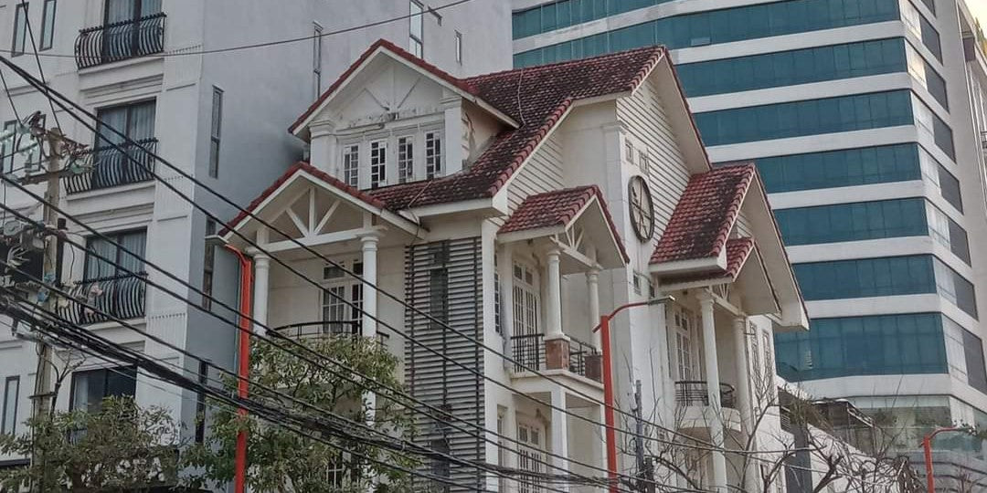 A traditional house with tall white pillars and a red roof sits nestled among two tall modern buildings in Da Nang, Vietnam