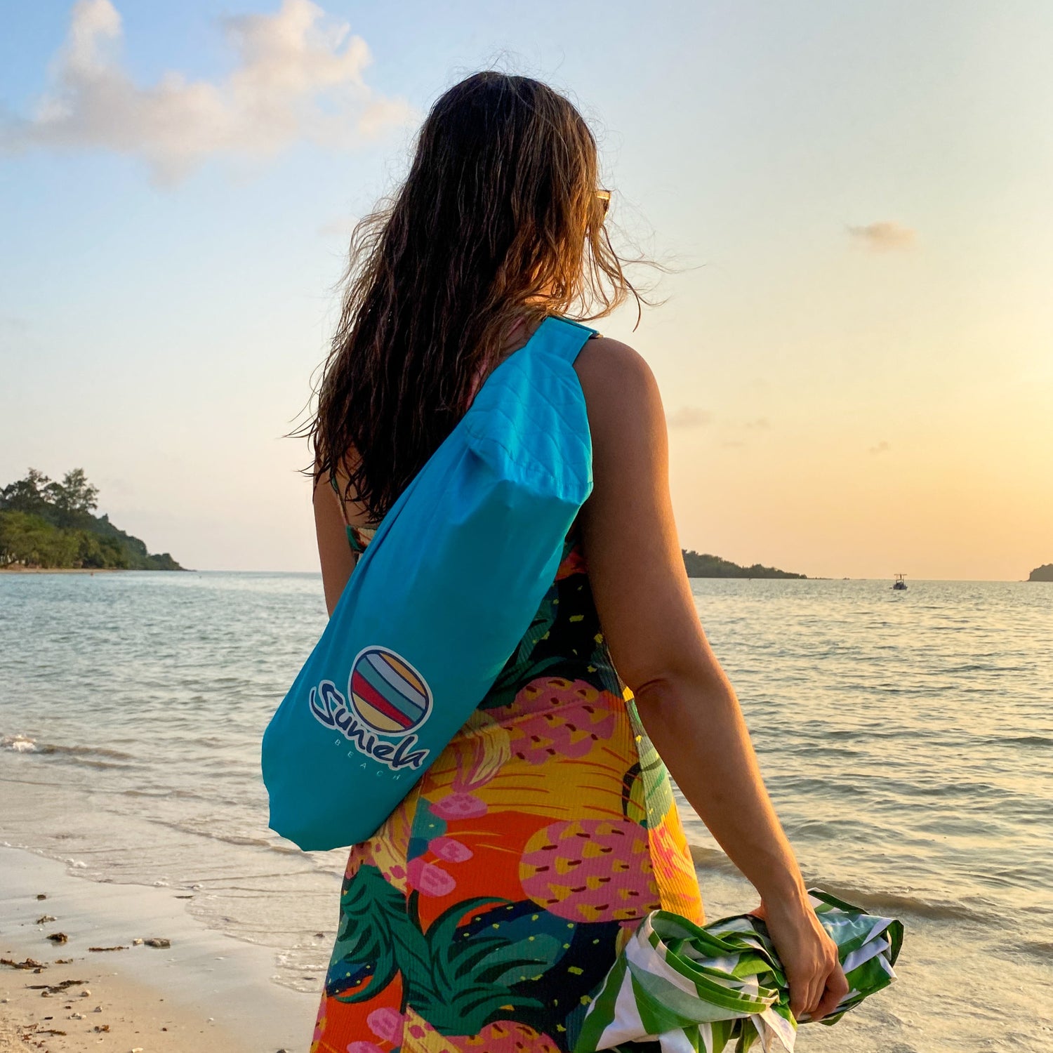 Woman wearing a colourful dress, carrying a Suniela shade cabana on her back. She's standing at the water's edge looking out at islands in the sunset_printTurquoiseSuniela