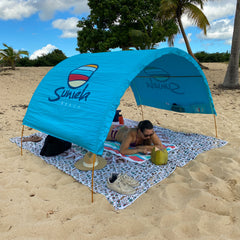 Person lying on a towel under a blue sun shelter with 'Sunela' branding on a sandy beach_printTurquoiseSuniela