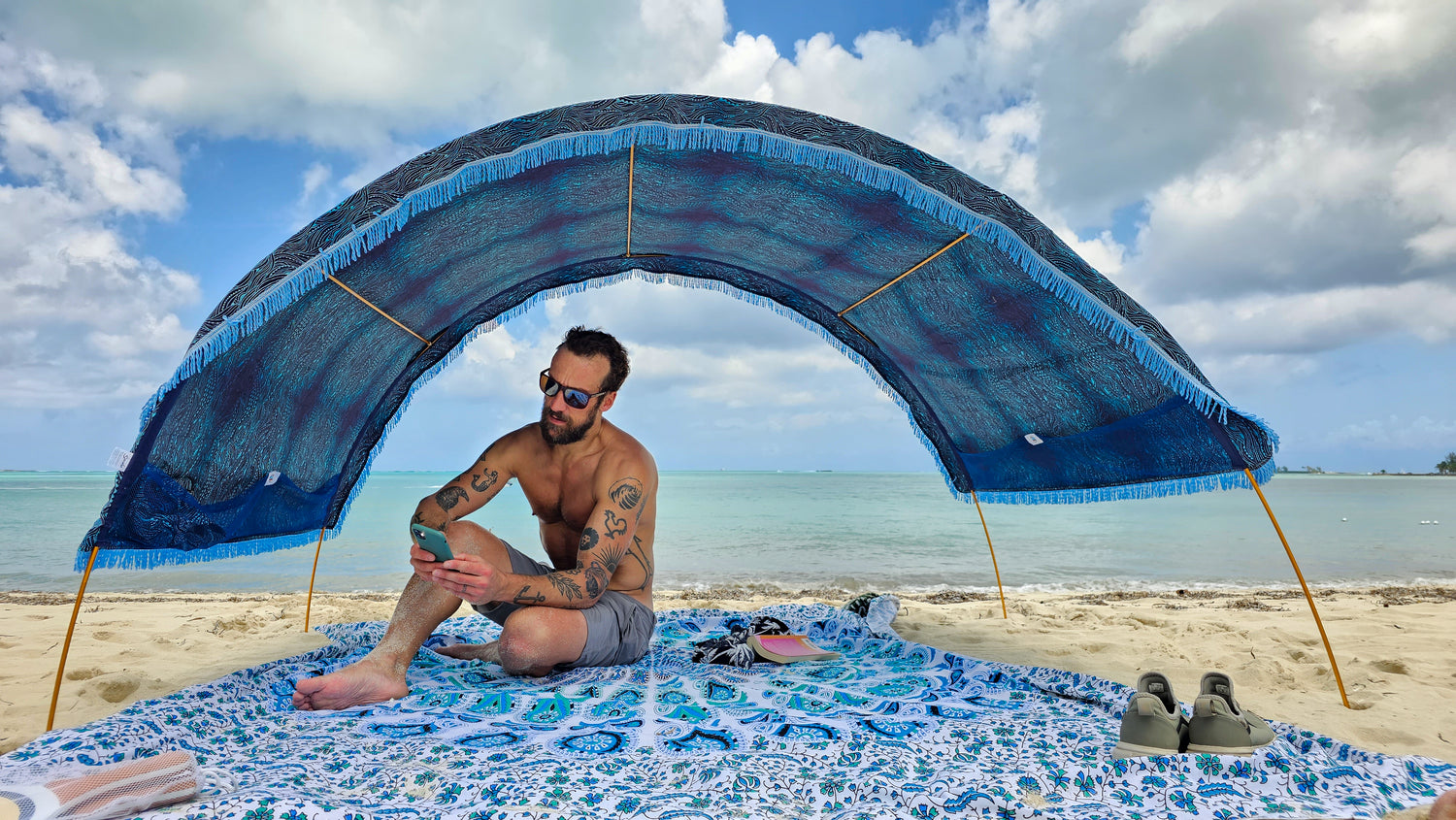 man sits on a tropical beach on his large Suniela Beach cotton mat in Blue Mandala pattern