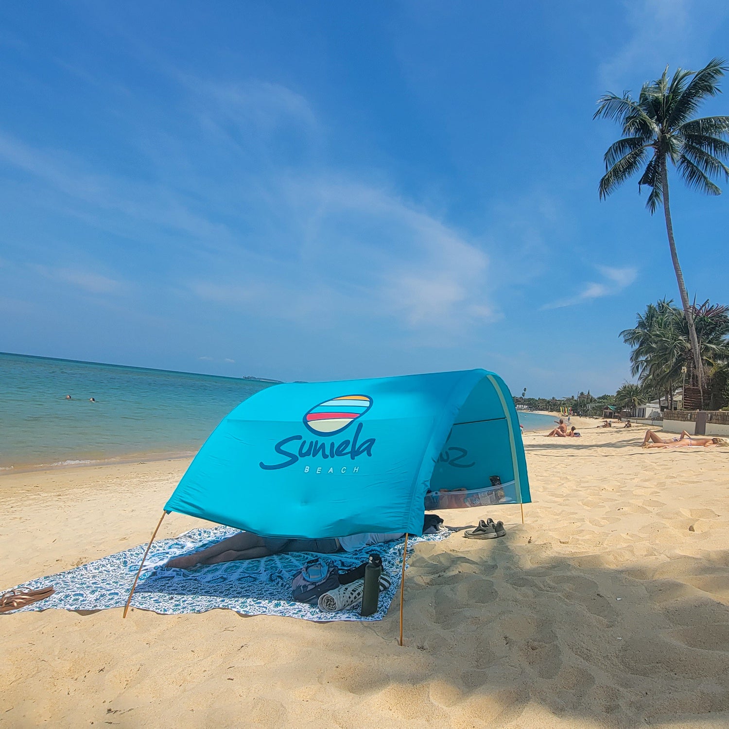 Blue beach tent with Suniela logo on a sandy beach with palm trees and ocean in the background